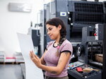 Eine Frau steht in einer Druckerei und prüft konzentriert einen großen Papierbogen vor einer Druckmaschine.