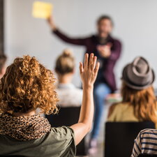 Eine Studierende meldet sich in einem Seminar, während vorne eine Person einen Vortrag hält.