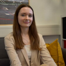 Eine junge Frau sitzt auf einem Sofa in einem Büro- oder Beratungsraum und blickt ruhig in die Kamera.