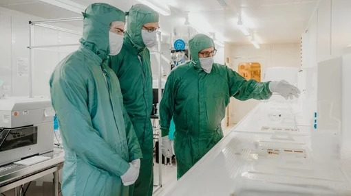 Drei Personen in Schutzkleidung arbeiten gemeinsam in einem Reinraum und begutachten technische Bauteile.