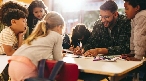 Ein Mann arbeitet gemeinsam mit mehreren Kindern an einem Tisch und unterstützt sie beim Zeichnen und Lernen.