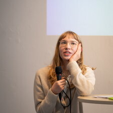 Eine junge Frau hält ein Mikrofon und spricht bei einer Veranstaltung oder Präsentation vor Publikum.