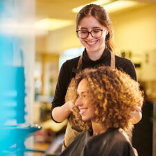 Eine Friseurin arbeitet lächelnd an den Haaren einer Kundin in einem Salon.