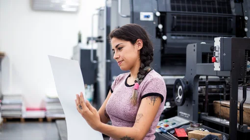 Eine Frau steht in einer Druckerei und prüft konzentriert einen großen Papierbogen vor einer Druckmaschine.