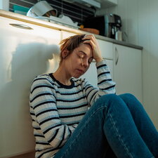 Frau sitzt auf dem Küchenboden an einen Schrank gelehnt, die Knie angezogen, eine Hand an der Stirn, die Augen geschlossen.