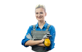 Frau in blauer Arbeitsbluse und grauer Latzhose hält Gehörschutz und Handschuhe und lächelt in die Kamera.