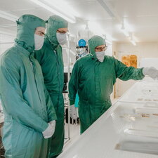 Drei Personen in Schutzkleidung arbeiten gemeinsam in einem Reinraum und begutachten technische Bauteile.