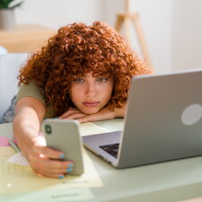 Eine Person mit lockigen Haaren sitzt zusammengesunken an einem Schreibtisch, legt den Kopf auf die Arme und hält in einer Hand ein Smartphone, während vor ihr ein geöffnetes Laptop und verstreute Unterlagen liegen.