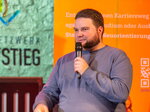 Ein Mann sitzt auf einer Bühne auf einem Hocker und spricht mit einem Mikrofon vor einem orangefarbenen Informationsbanner.