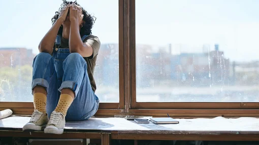Eine junge Person sitzt nachdenklich am Fenster, hält den Kopf in den Händen; neben ihr liegen ein Smartphone und ein Notizbuch.