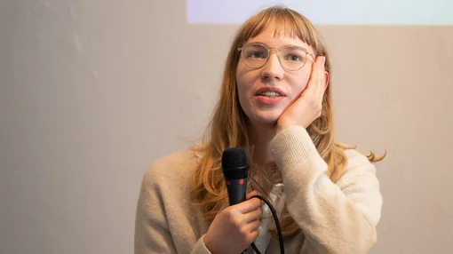 Eine junge Frau hält ein Mikrofon und spricht bei einer Veranstaltung oder Präsentation vor Publikum.