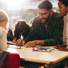 Ein Mann arbeitet gemeinsam mit mehreren Kindern an einem Tisch und unterstützt sie beim Zeichnen und Lernen.