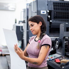 Eine Frau steht in einer Druckerei und prüft konzentriert einen großen Papierbogen vor einer Druckmaschine.