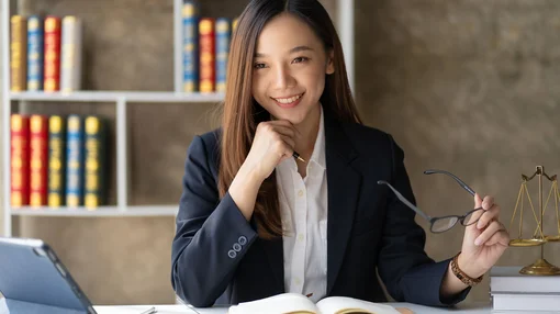 Eine Frau sitzt an einem Schreibtisch mit Buch und Tablet, hält eine Brille und lächelt in einem Büro- oder Studienumfeld.