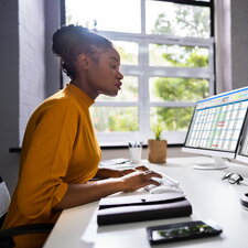 Eine Frau arbeitet konzentriert an einem Schreibtisch mit zwei Monitoren, auf denen Tabellen und Daten angezeigt werden.