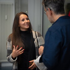 Zwei Personen unterhalten sich in einem hellen Raum mit Heizkörper und Tür im Hintergrund. Eine junge Frau mit langen braunen Haaren gestikuliert lebhaft; ein älterer Mann mit Brille und blauem Hemd hört aufmerksam.