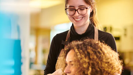Eine Friseurin arbeitet lächelnd an den Haaren einer Kundin in einem Salon.