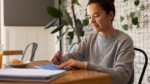 Eine junge Frau sitzt an einem Tisch, arbeitet konzentriert mit Unterlagen und schreibt Notizen in ruhiger Umgebung.