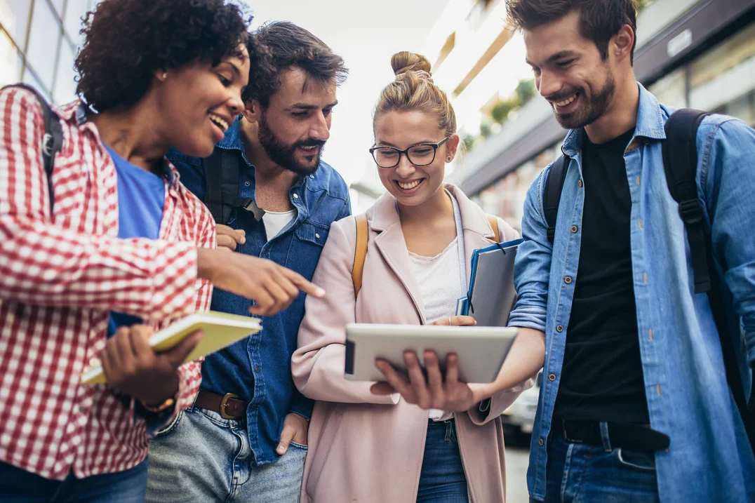 Four young adults stand together outdoors, smiling as they look at a tablet held by one of them; they appear engaged in conversation and are carrying backpacks and notebooks.