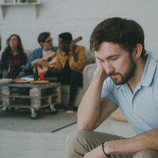Nachdenklicher junger Mann sitzt im Vordergrund, während im Hintergrund eine Gruppe zusammen sitzt und Musik macht.