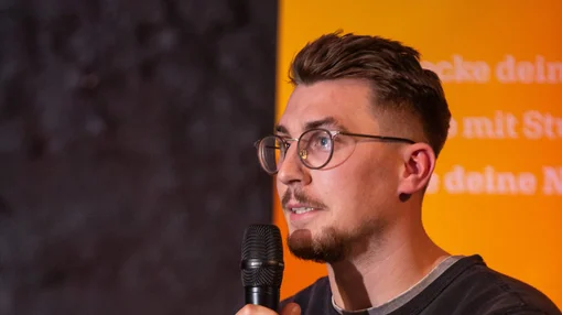 Ein Mann mit Brille hält ein Mikrofon und spricht bei einer Veranstaltung vor einer Präsentationswand.