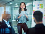 Eine junge Frau präsentiert vor einem Team in einem modernen Büro und erklärt Inhalte an einem Whiteboard mit Haftnotizen.