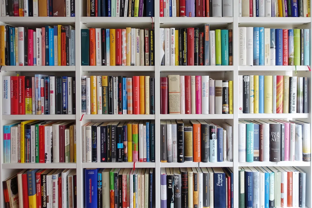 Bunte Bücher stehen eng gedrängt in einem weißen Bücherregal.