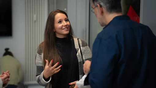 Zwei Personen unterhalten sich in einem hellen Raum mit Heizkörper und Tür im Hintergrund. Eine junge Frau mit langen braunen Haaren gestikuliert lebhaft; ein älterer Mann mit Brille und blauem Hemd hört aufmerksam.