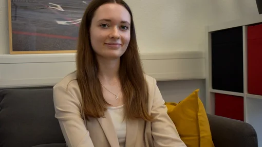 Eine junge Frau sitzt auf einem Sofa in einem Büro- oder Beratungsraum und blickt ruhig in die Kamera.