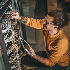 Ein Mann arbeitet an einem Serverschrank, überprüft Kabelverbindungen und hält ein Tablet in der Hand.