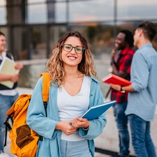 Eine Studierende steht mit Notizbuch und Rucksack vor einem Hochschulgebäude, im Hintergrund unterhalten sich weitere Studierende.