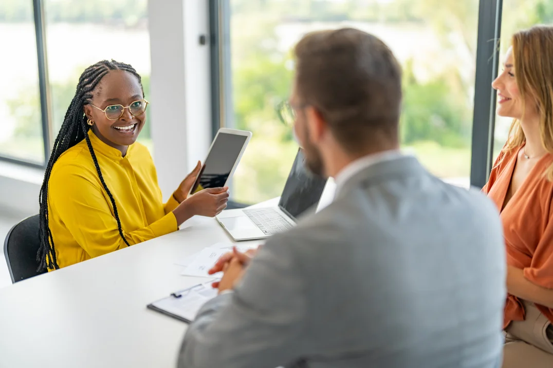 Drei diverse Personen sitzen an einem Konferenztisch mit Laptops und Papieren: Eine lachende Frau mit Brille und Zöpfen hält ein Tablet hoch, ein Mann mit Brille und eine Frau lächeln zu.