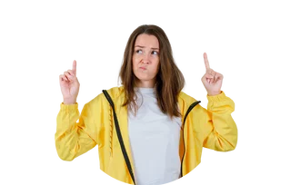 Junge Frau mit braunen Haaren und gelbem Regenmantel zeigt mit beiden Zeigefingern nach oben und blickt dabei fragend.