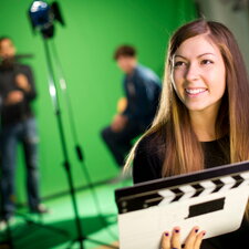 Eine Studierende hält eine Filmklappe in einem Medien- oder Filmstudio vor einem Greenscreen.