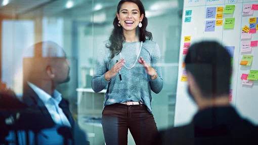 Eine junge Frau präsentiert vor einem Team in einem modernen Büro und erklärt Inhalte an einem Whiteboard mit Haftnotizen.