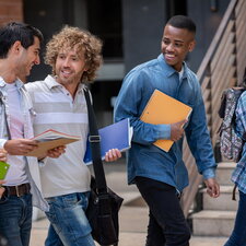 Eine Gruppe junger Erwachsener geht lächelnd gemeinsam über einen Campus und trägt Hefte, Notizbücher und Rucksäcke.