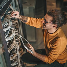 Ein Mann arbeitet an einem Serverschrank, überprüft Kabelverbindungen und hält ein Tablet in der Hand.
