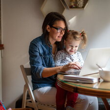 Eine Frau arbeitet an einem Laptop, während ein kleines Kind neben ihr sitzt und zuschaut, in einem häuslichen Umfeld.