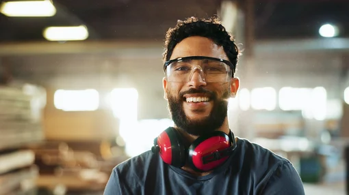 Ein Mann mit Schutzbrille und Gehörschutz steht lächelnd in einer Werkstatt oder Produktionshalle.