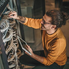 Ein Mann arbeitet an einem Serverschrank, überprüft Kabelverbindungen und hält ein Tablet in der Hand.