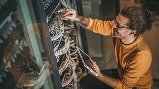 Ein Mann arbeitet an einem Serverschrank, überprüft Kabelverbindungen und hält ein Tablet in der Hand.