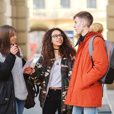 Drei junge Studierende stehen draußen vor einem Gebäude und unterhalten sich; sie halten Notizbücher und tragen Winterjacken und Rucksäcke.