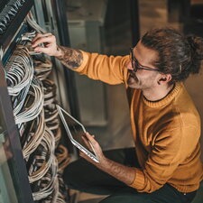 Ein Mann arbeitet an einem Serverschrank, überprüft Kabelverbindungen und hält ein Tablet in der Hand.