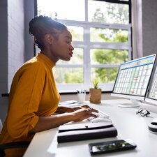 Eine Frau arbeitet konzentriert an einem Schreibtisch mit zwei Monitoren, auf denen Tabellen und Daten angezeigt werden.