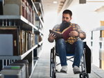 Student im Rollstuhl sitzt in einem Bibliotheksgang und liest ein großes Buch, umgeben von Bücherregalen.