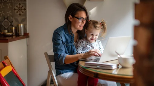 Eine Frau arbeitet an einem Laptop, während ein kleines Kind neben ihr sitzt und zuschaut, in einem häuslichen Umfeld.