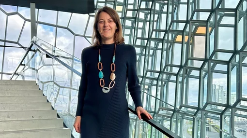 Eine Frau steht auf einer Treppe in einem modernen Glasgebäude und lächelt in die Kamera.