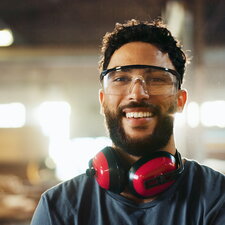 Ein Mann mit Schutzbrille und Gehörschutz steht lächelnd in einer Werkstatt oder Produktionshalle.