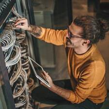 Ein Mann arbeitet an einem Serverschrank, überprüft Kabelverbindungen und hält ein Tablet in der Hand.