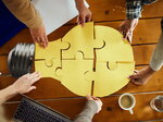 Vier Hände von verschiedenen Personen greifen von oben in eine große gelbe Puzzel-Lampe (Glühbirne), die aus Puzzleteilen besteht, auf einem Holztisch mit Laptop und Tassen.